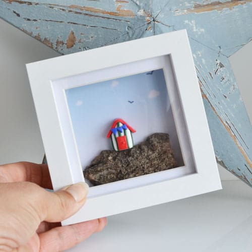 Miniature clay beach hut set on driftwood and framed in a white square frame.