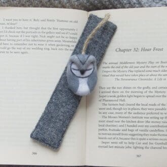 A bookmark made from blue/grey wool tweed with a needle felted grey and white owl attached from the top by a jute cord. The owl has it's eyes closed and is resting on an open book.