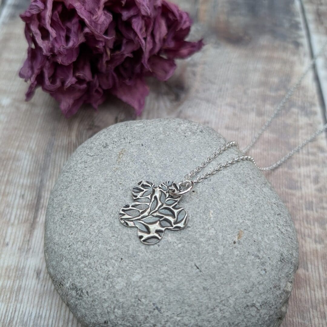 An oxidized silver pendant with a floral leaf pattern design on a smooth gray stone background. The pendant is on a silver chain.