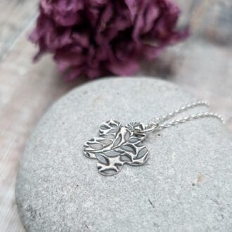 An oxidized silver pendant with a floral leaf pattern design on a smooth gray stone background. The pendant is on a silver chain.