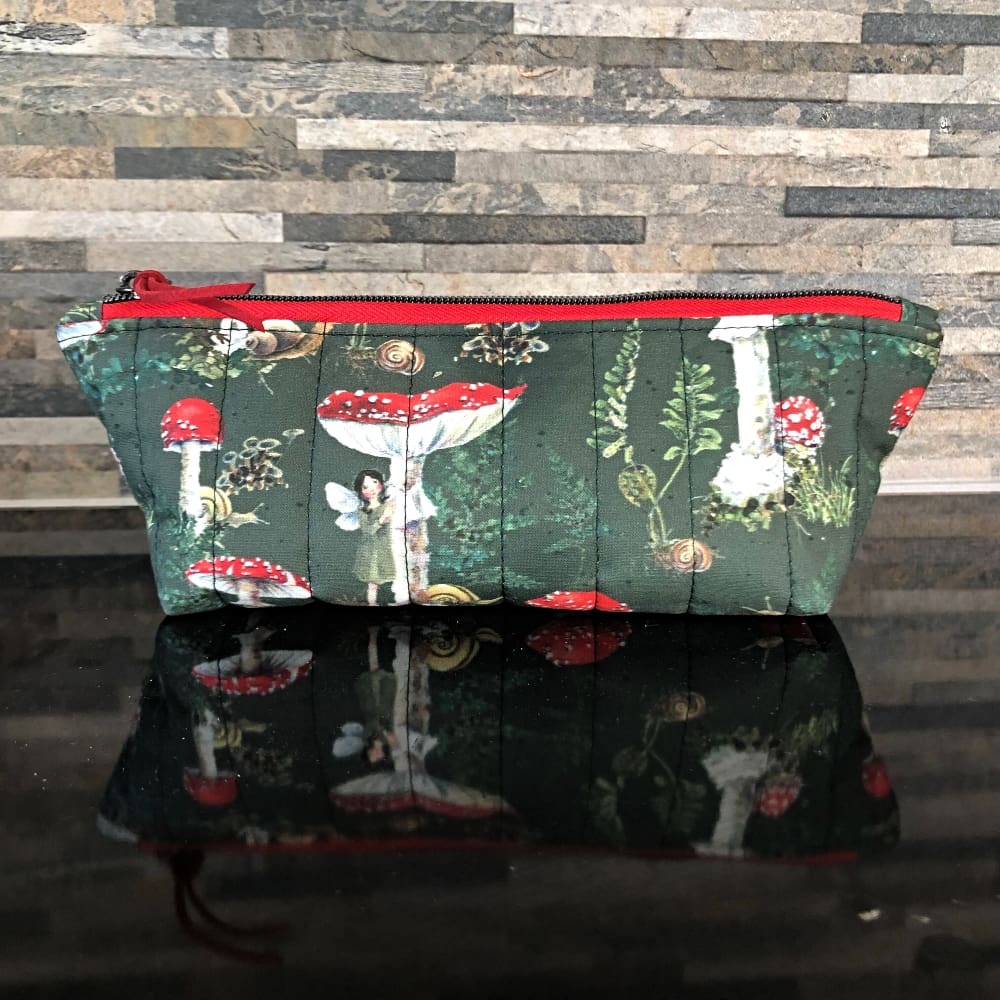 Pencil case or make up bag in dark green woodland fabric featuring fly algaric mushrooms and whimsical woodland folk, red zip and pale green lining.