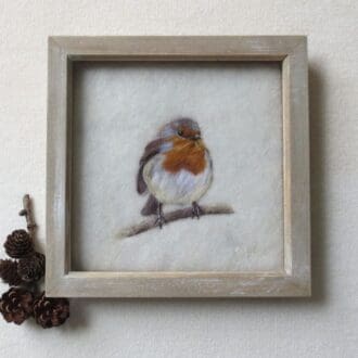 A handmade needle felted wool picture of a robin on a cream wool background and in a pale wood box frame. The robin is facing forward and sat on a branch.