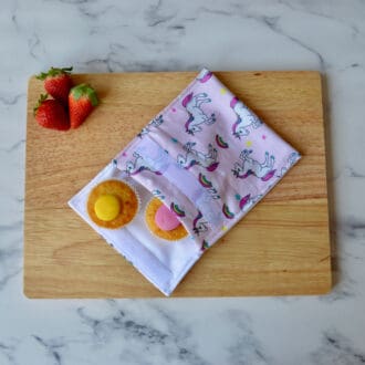 Light pink unicorn snack bag on a wooden board with cupcakes inside and fresh strawberries.