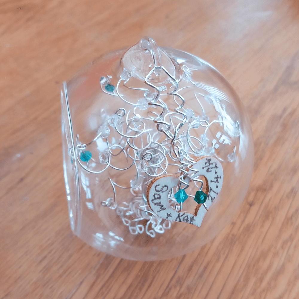 8cm glass bauble with a wooden heart charm that is attached to the side of the bauble with a silver wire. Names and a date are handwritten on, and it's decorated with an emerald and blue zircon drops.