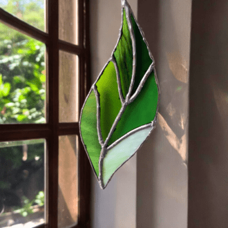 a leaf-shaped piece of stained glass with various shades of green and white, outlined with silver solder lines.