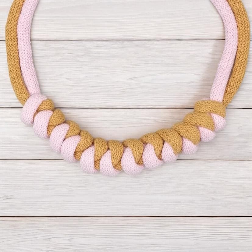 Close up of knot feature on chunky modern pink and mustard rope necklace, shown against a light wooden worktop background.