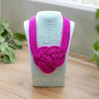 Pink chunky 4 strand knotted statement necklace with feature knot in center displayed on a stand atop a light wood coutnertop.