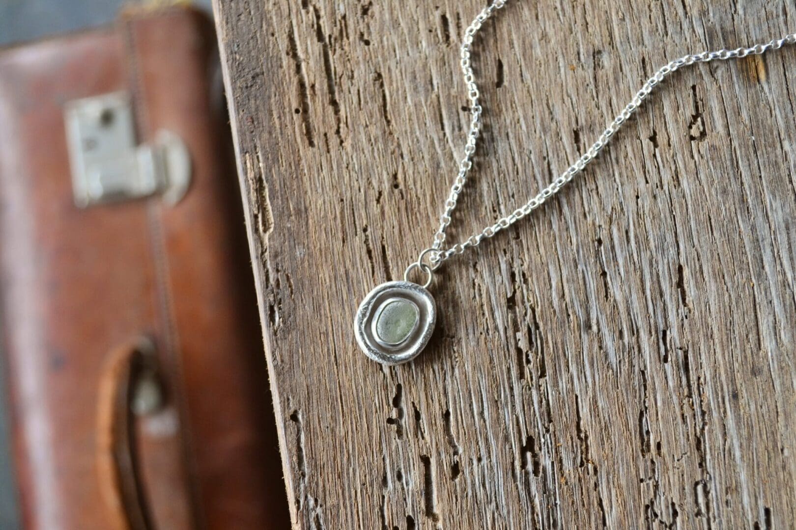 Pale green sea glass surrounded by molten silver. The pendant is shown on a silver chain. The necklace lays on an old wooden tabletop. a vintage leather suitcase is shown below