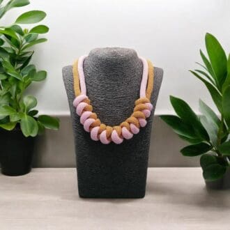 Pink and mustard chunky modern necklace shown on a model bust on a light woodne worktop with plants in the background.