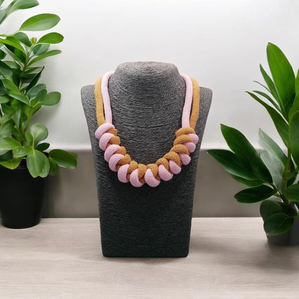 Pink and mustard chunky modern necklace shown on a model bust on a light woodne worktop with plants in the background.