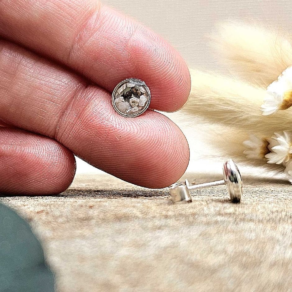 Close-up of jeweller's fingers showcasing small round handmade silver keepsake stud earrings with ashes set in clear resin