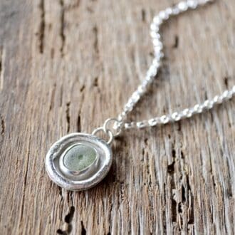 A light green sea glass pendant, with molten silver surround is strung on a silver chain. The necklace lays on an old wooden surface