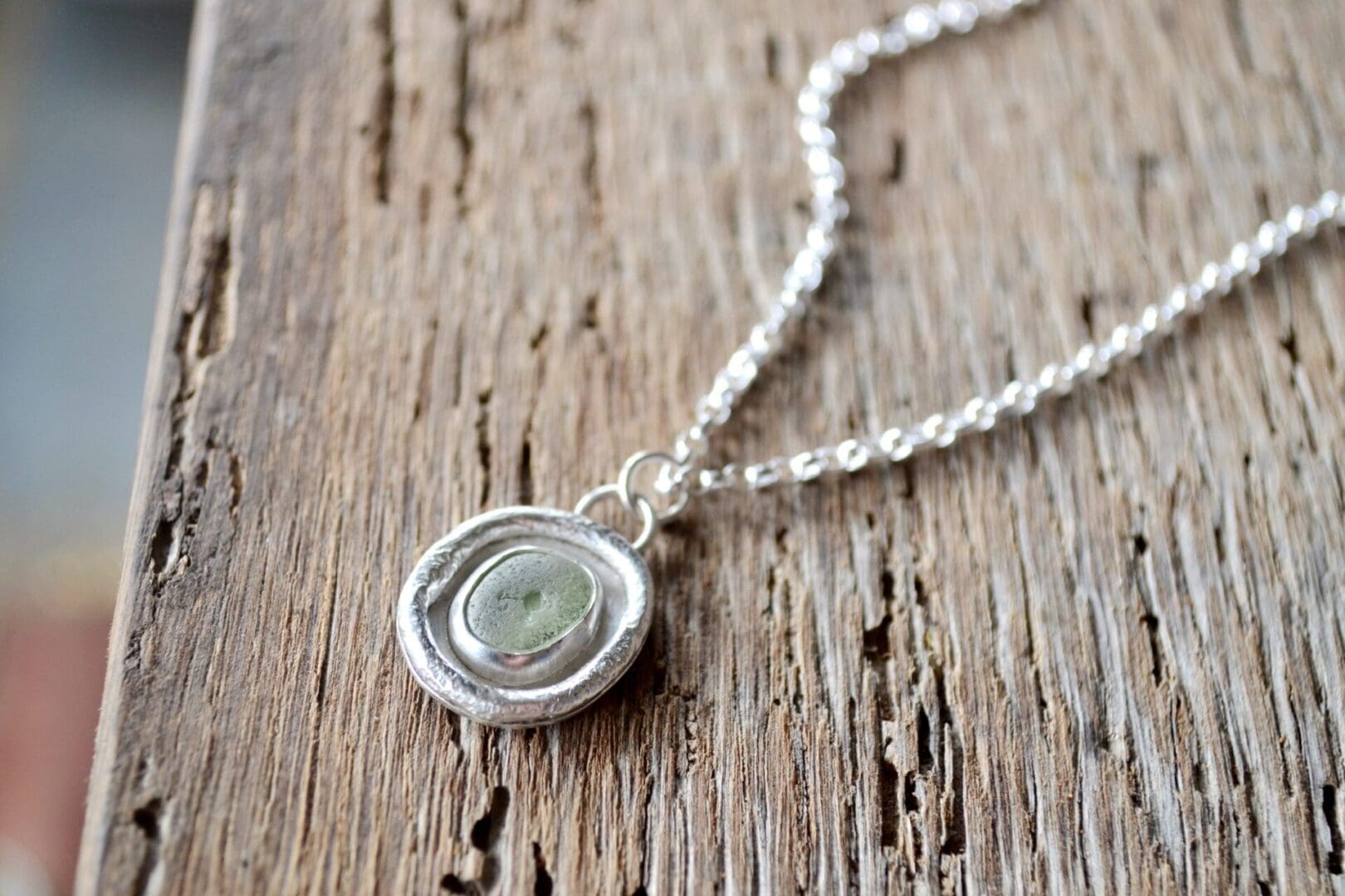 A light green sea glass pendant, with molten silver surround is strung on a silver chain. The necklace lays on an old wooden surface