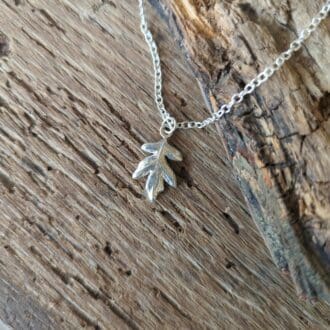 A silver hawthorn leaf pendant on a silver chain lays on textured wood