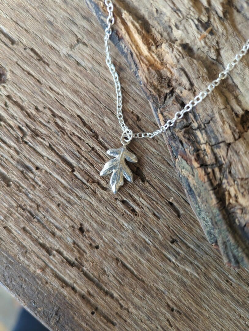 A silver hawthorn leaf pendant on a silver chain lays on textured wood