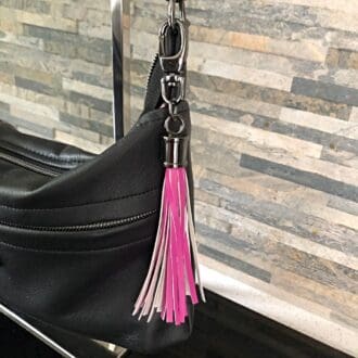 A pink faux leather fringed bag charm or tassel hanging from a black bag against a narrow brick wall