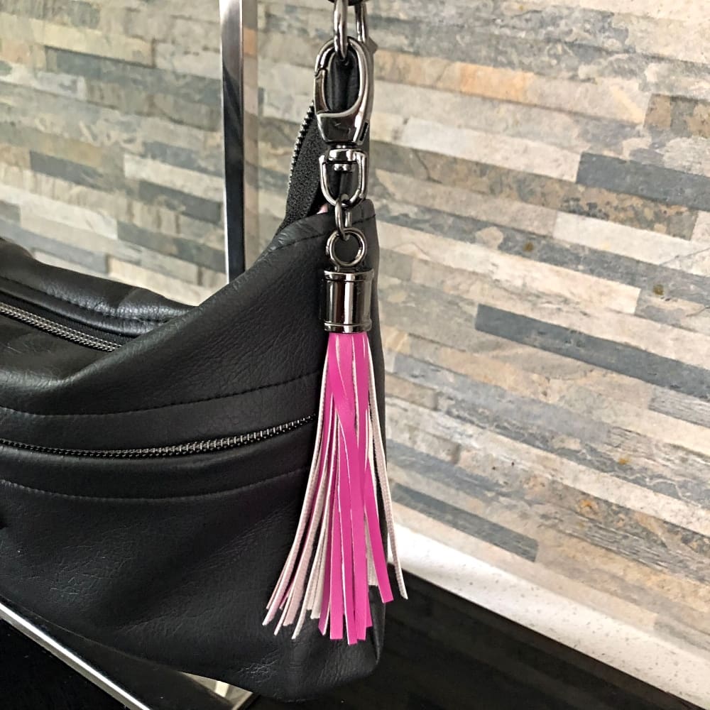 A pink faux leather fringed bag charm or tassel hanging from a black bag against a narrow brick wall