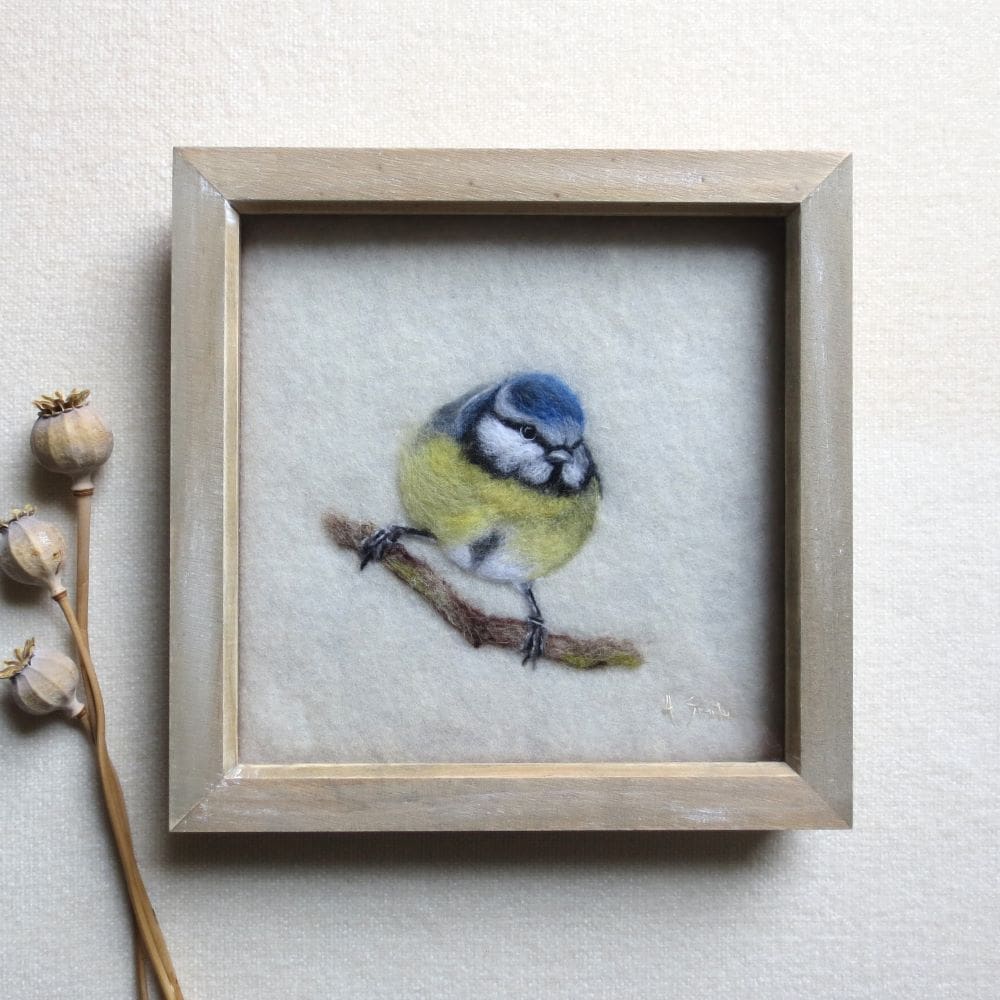 A handmade needle felted wool painting of a blue-tit on a cream wool background and in a pale wood box frame. The bird is sat on a branch and facing forward.