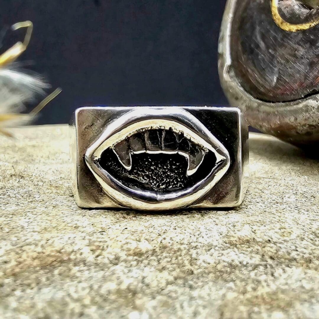 Handcrafted recycled sterling silver vampire fanged mouth signet ring displayed on decorative stone next to victorian bobbin