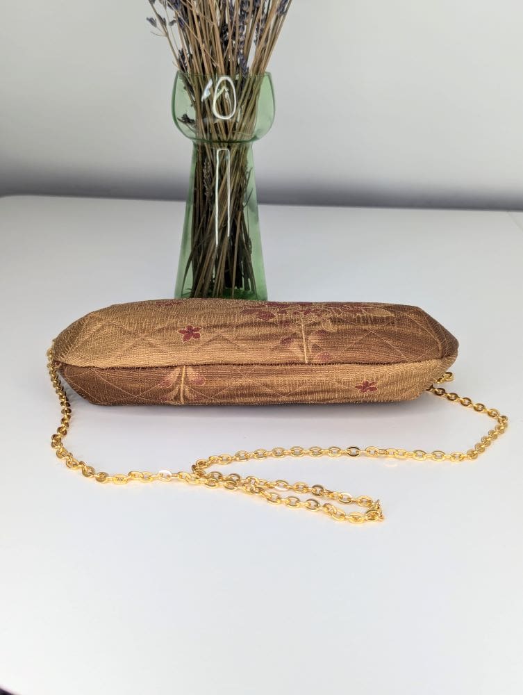 A gold coloured handmade shoulder bag laying on its side showing the underside view with a gold coloured copper chain