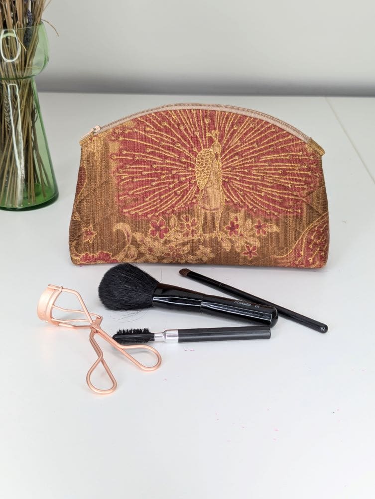 A handmade make up pouch with peacock design on one side. It has a zip fastening and is shown on a white surface with make up brushes in front