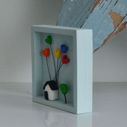 A miniature wooden frame with a rustic or distressed finish with a small clay house with coloured hearts inside.