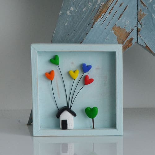 A miniature wooden frame with a rustic or distressed finish with a small clay house with coloured hearts inside.