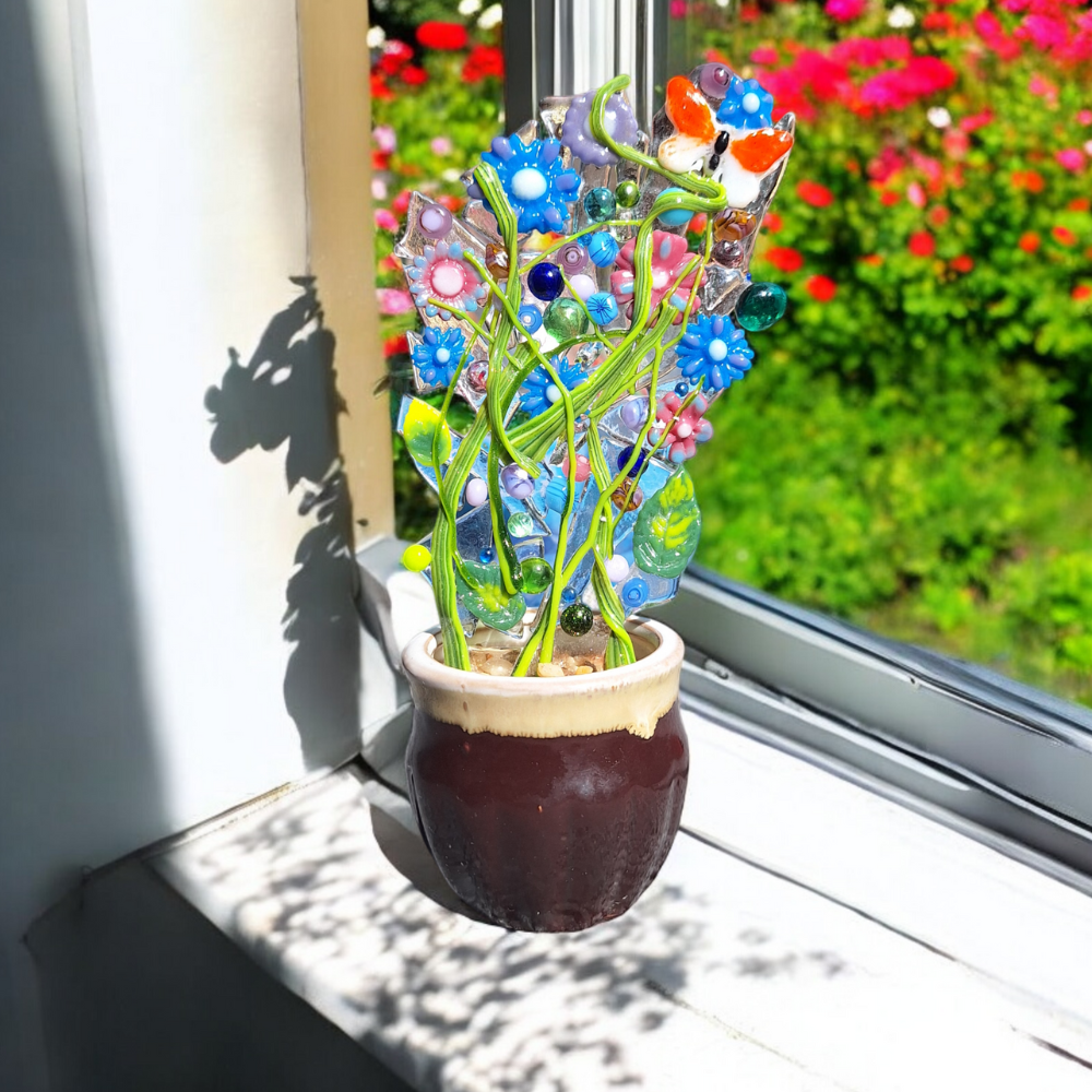 A vibrant fused glass bouquet ornament sits in a small ceramic pot on a windowsill, with colourful garden flowers visible in the background. The glass sculpture features a variety of bright colours, including blue, green, and red, with intricate floral and leaf designs. Sunlight streams through the window, casting a decorative shadow on the wall and illuminating the glass bouquet, enhancing its vibrant colours and artistic details.