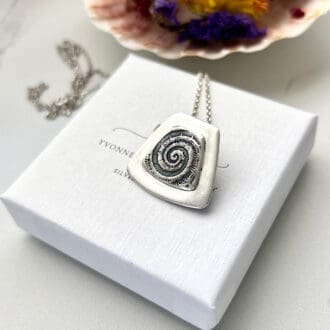 A square silver pendant with a fossil embossed on the front resting on a small white gift box.