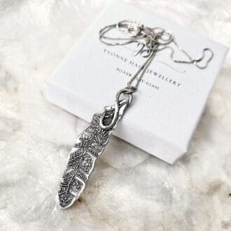 A silver pendant in the shape of a feather sat on a white gift box.