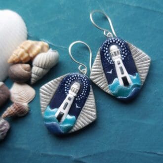 A pair of handmade polymer clay earrings depicting an Art Deco style white lighthouse emerging from wavesblue and turquoise waves, against a dark blue sky. The image shows the Art Deco style, blue, white and grey colouration and Sterling silver handmade ear wires.