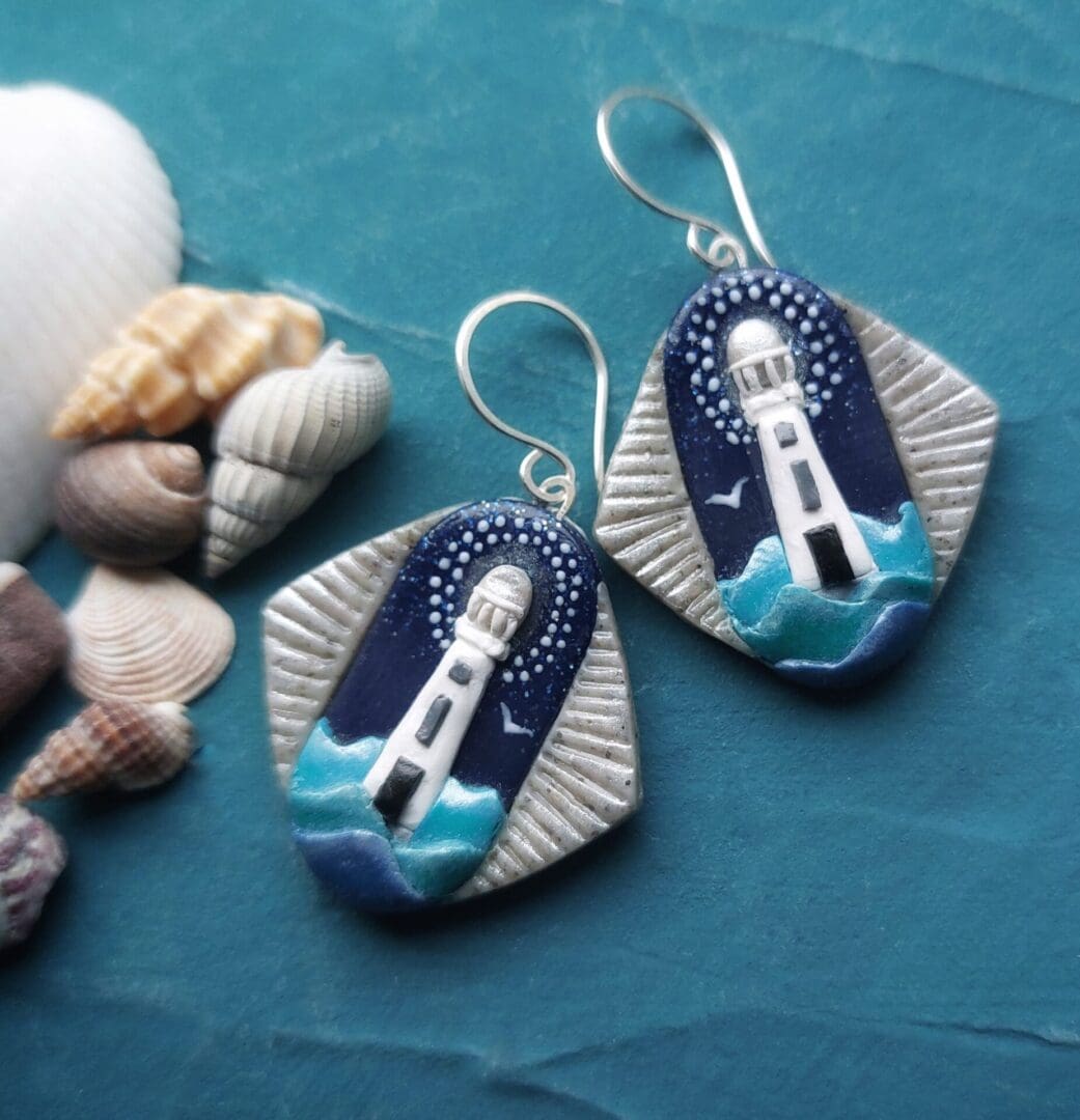 A pair of handmade polymer clay earrings depicting an Art Deco style white lighthouse emerging from wavesblue and turquoise waves, against a dark blue sky. The image shows the Art Deco style, blue, white and grey colouration and Sterling silver handmade ear wires.