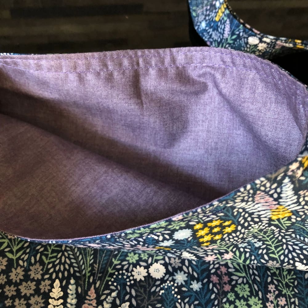 Adjustable, cross body style mastectomy drain bag in a floral fabric of stylised yellow white delphiniums, on a navy blue background and lilac cotton lining, lying on a reflective black surface.