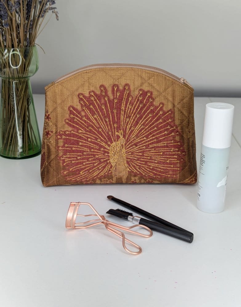 A handmade make up pouch. Gold and red in colour with a peacock design on one side. Shown on a white surface with make up in front and to the side