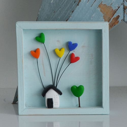 A miniature wooden frame with a rustic or distressed finish with a small clay house with coloured hearts inside.