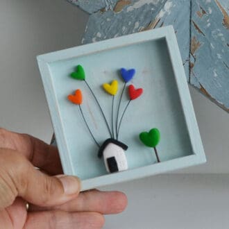 A miniature wooden frame with a rustic or distressed finish with a small clay house with coloured hearts inside.