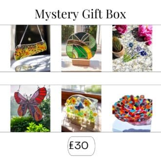 Collage image titled 'Mystery Gift Box' showcasing a variety of fused glass art pieces. The top row includes a floral glass hanging, a green and yellow stained glass panel on a wooden stand, and three glass flower stakes. The bottom row features a red and orange butterfly suncatcher, a decorative glass arch with embedded flowers, and a colourful, multi-hued glass bowl. Price displayed at the bottom: £30."