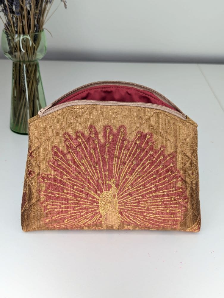 A handmade, quilted, gold make up pouch with a peacock design. It has a zip closure and is open showing the red lining