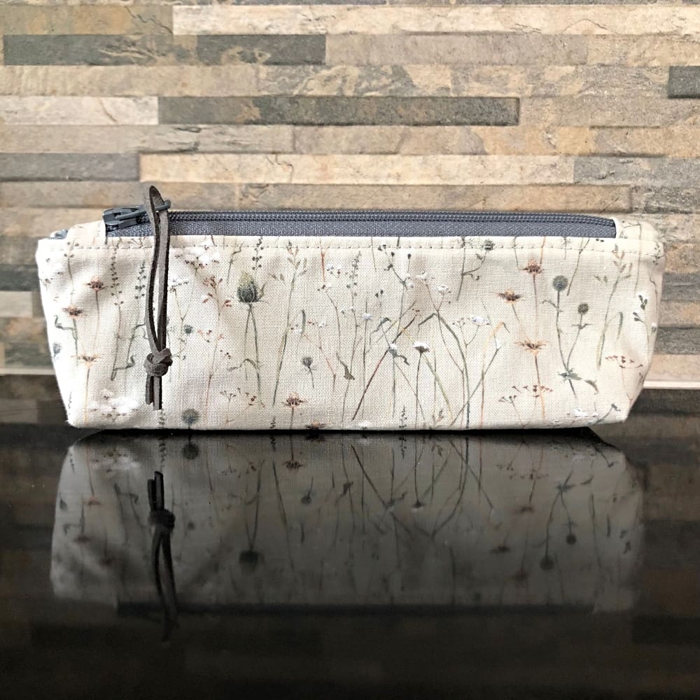 Pencil case or make up bag in pale grey-blue fabric featuring winter grasses and seedheads, with a dark grey zip, faux suede pull and lining