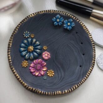 A round black trinket dish with floral decorations in pink, purple and blue with gold details. The dish comes with a pair of blue and gold flower stud earrings and has holes for storage of additional pairs of studs. Handmade in polymer clay by Weaversfield Jewellery on The British Craft House.