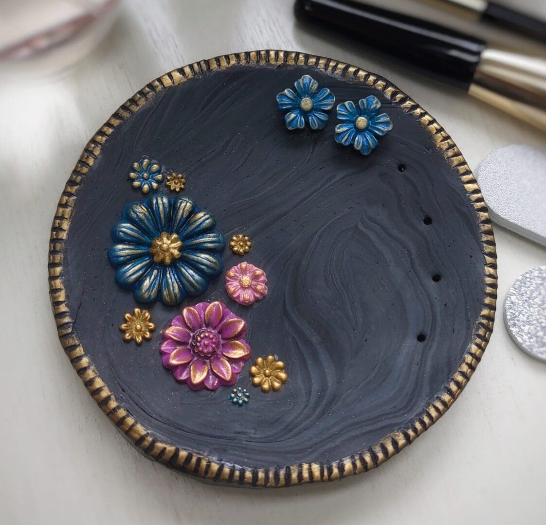 A round black trinket dish with floral decorations in pink, purple and blue with gold details. The dish comes with a pair of blue and gold flower stud earrings and has holes for storage of additional pairs of studs. Handmade in polymer clay by Weaversfield Jewellery on The British Craft House.