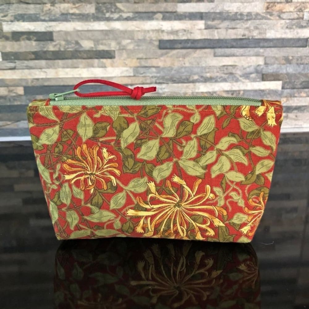 A small sized zipped make up bag in William Morris Honeysuckle fabric in gold on a rich red background. It has a pale green zip and lining.