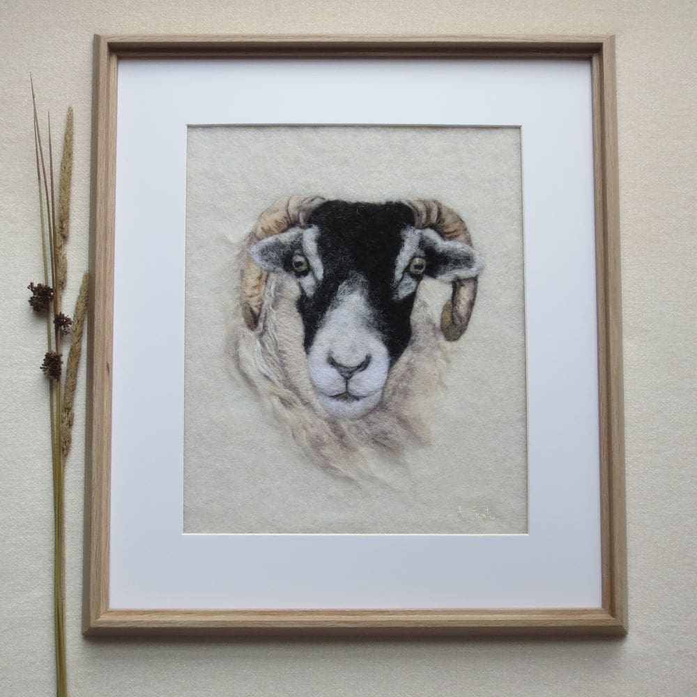 A handmade wool painting of the face of a swaledale sheep on a cream wool background. The sheep is facing forward and is in a pale wood frame with a cream coloured mount.