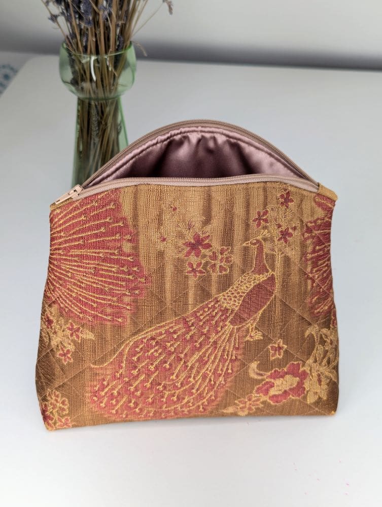 A gold and red toiletry bag with a peacock design, a zip opening showing the polyester satin lining