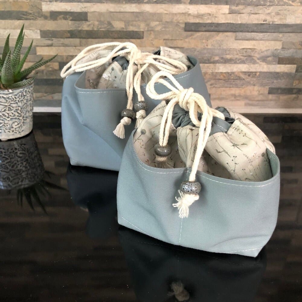 Two large drawstring bags in grey-blue fabric with winter grasses and seedheads. Full width external pocket options in solid blue-grey fabric. They close with natural unbleached cotton secured with chunky ceramic beads on a reflective black surface. In small or large