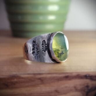 Moss agate cabochon set in wide sterling silver ring band featuring pine tree motifs.