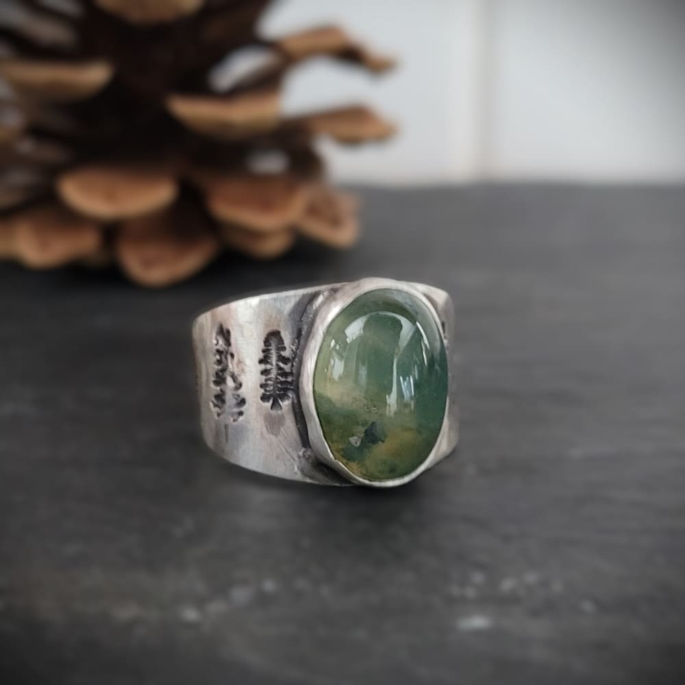 Moss agate cabochon set in wide sterling silver ring band featuring pine tree motifs.