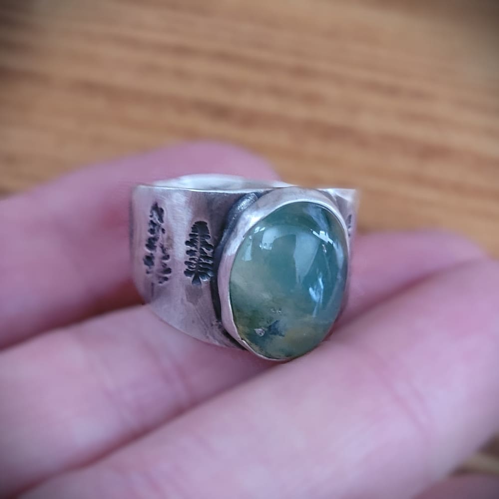 Moss agate cabochon set in wide sterling silver ring band featuring pine tree motifs.