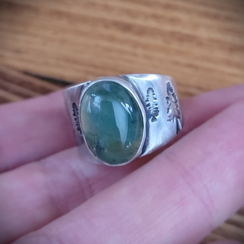 Moss agate cabochon set in wide sterling silver ring band featuring pine tree motifs.