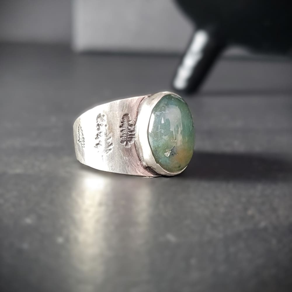 Moss agate cabochon set in wide sterling silver ring band featuring pine tree motifs.